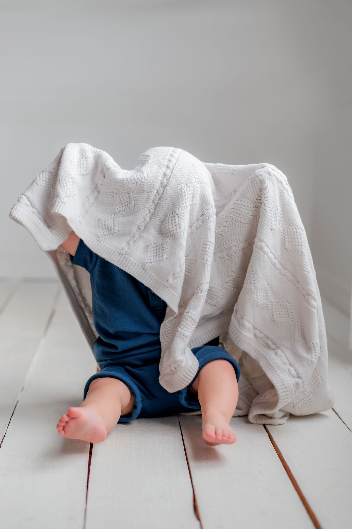 Small bare feet peek out from under a white blanket and blue pants on a light wood floor.