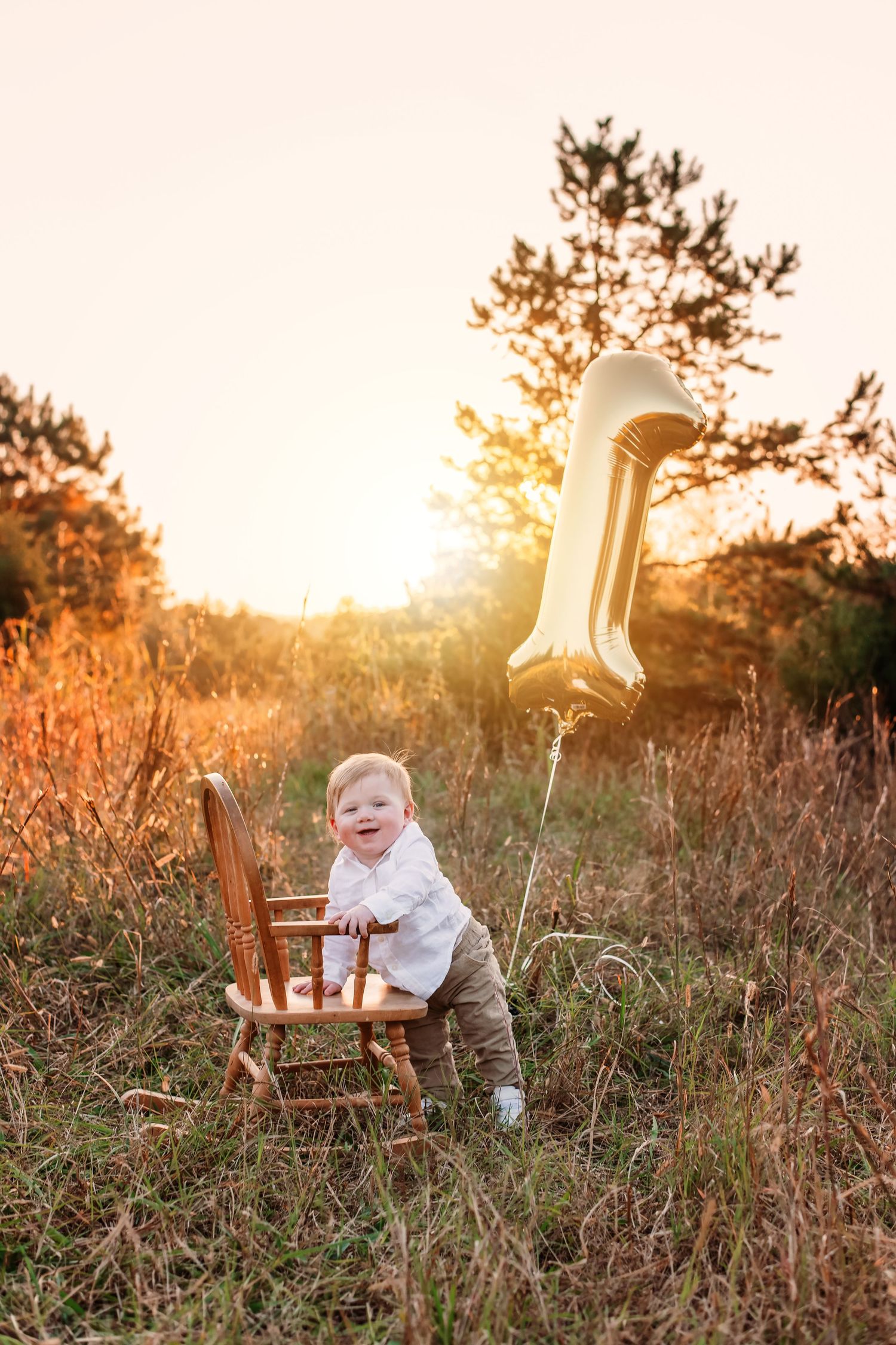 Baby celebrates first birthday with wooden chair and gold balloon in sunset field.