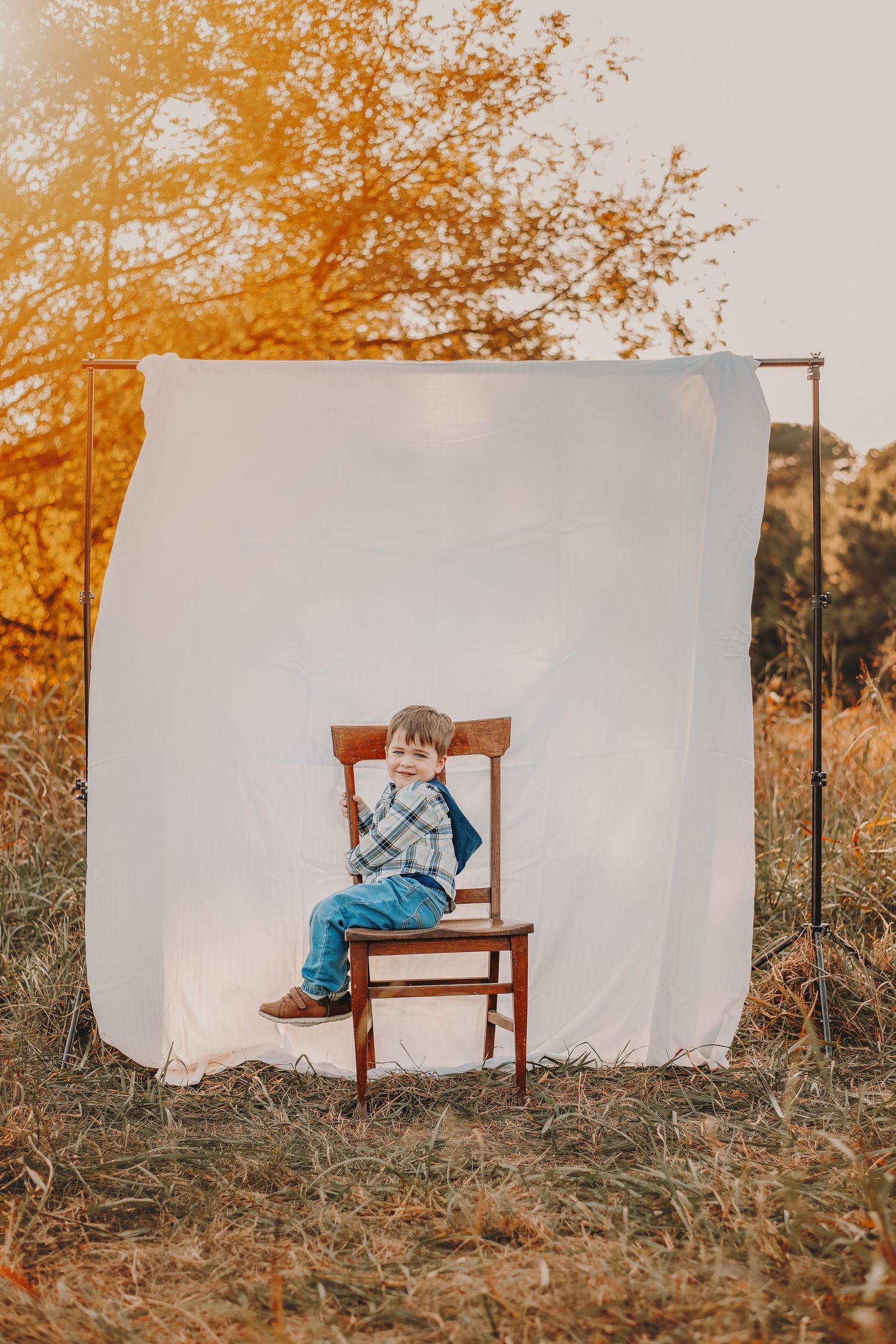 A white sheet backdrop hangs in a golden autumn field with a wooden chair, creating a rustic outdoor photo setup.