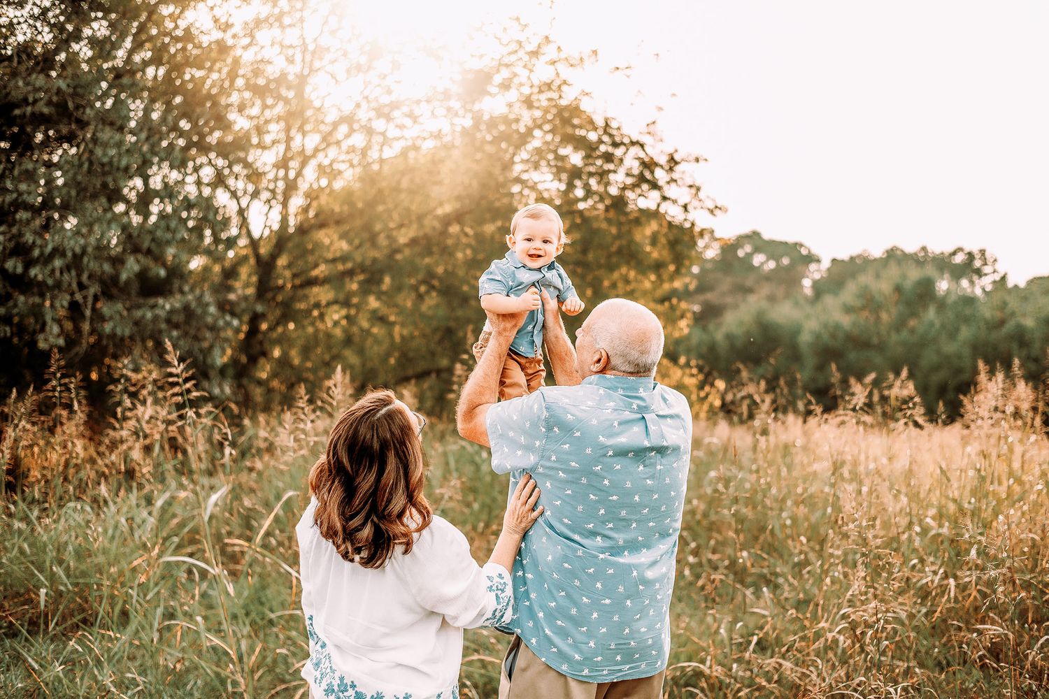 Parents lifting a laughing baby in a sunlit meadow during a golden hour family photo session outdoors.