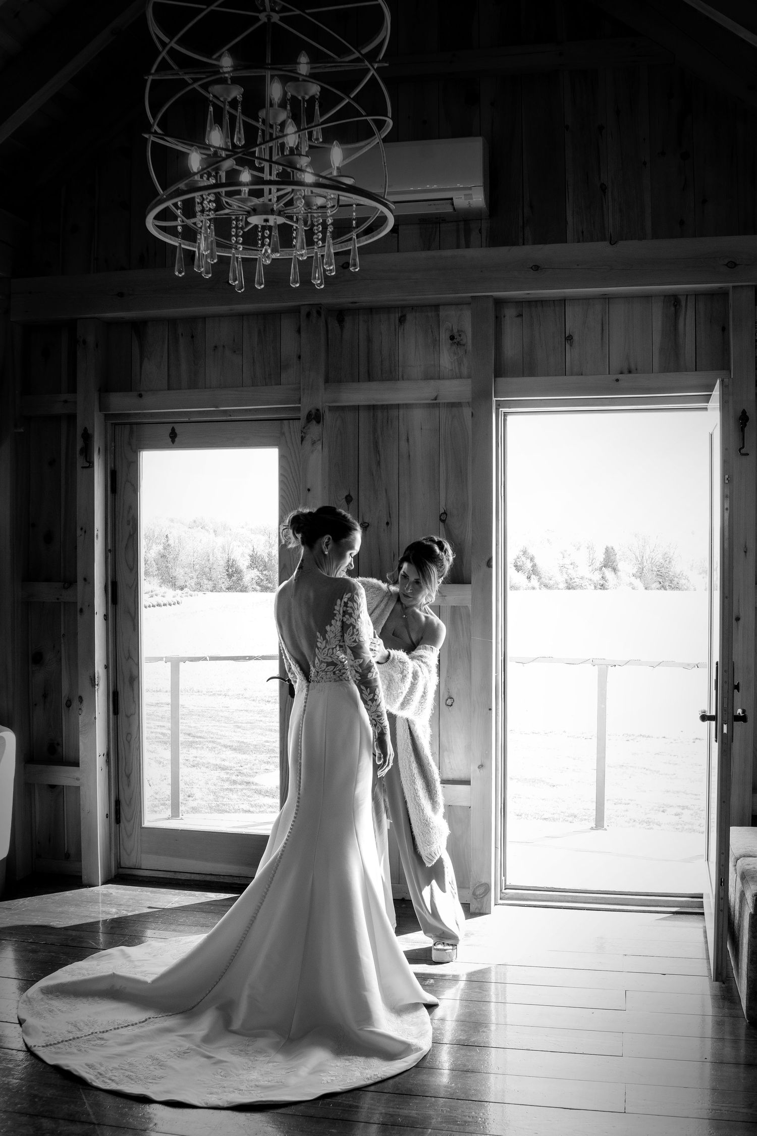 Bride in lace wedding gown with train stands by open barn doors in black and white photo.