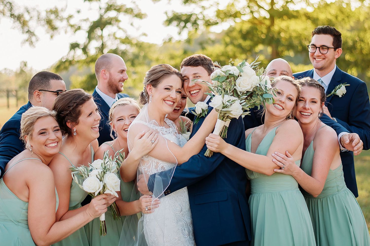 Joyful wedding party in sage green bridesmaid dresses and navy suits celebrating with the smiling bride and groom outdoors.