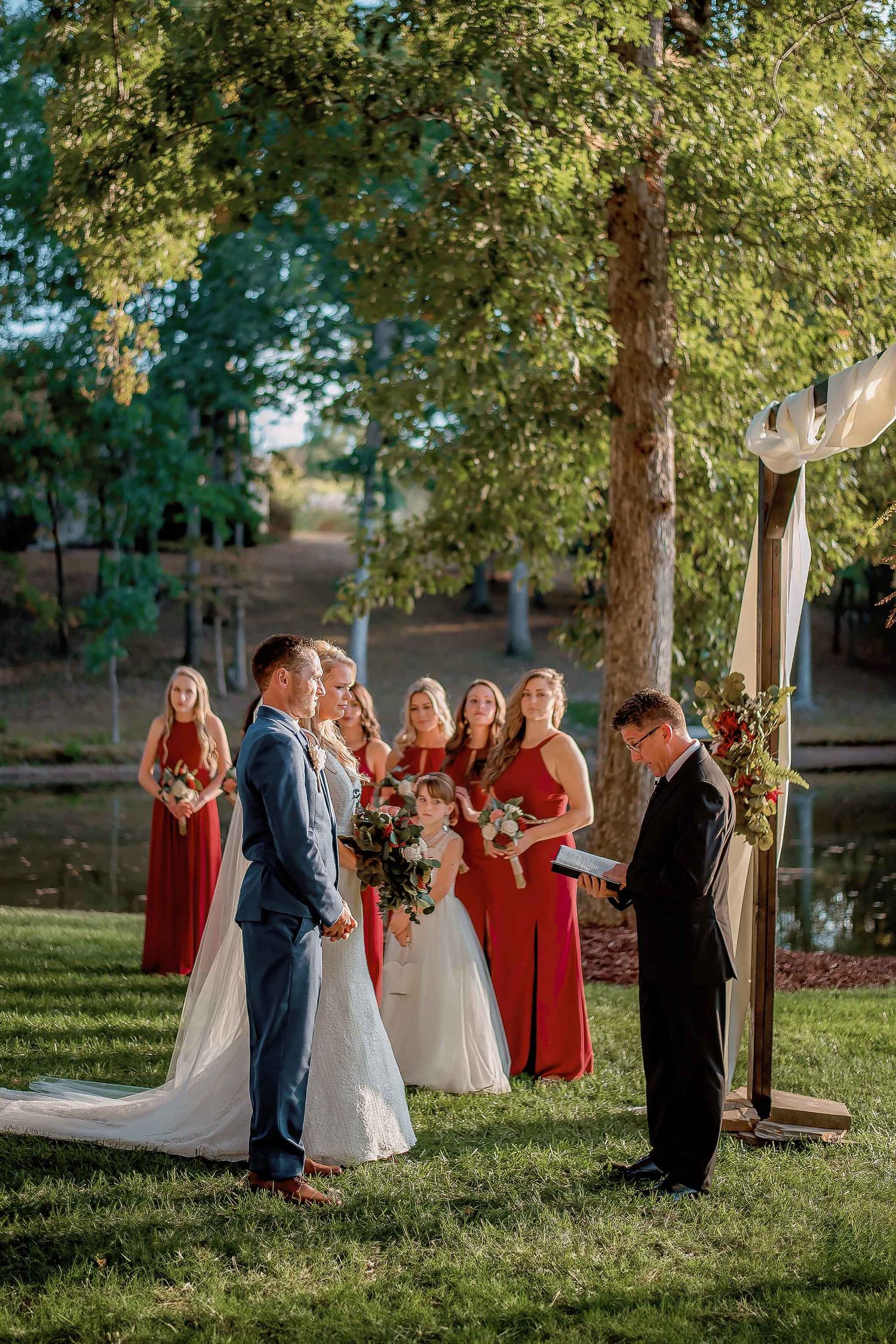 Outdoor wedding ceremony with bride, groom, officiant, and red-dressed bridesmaids by a pond under lush green trees.