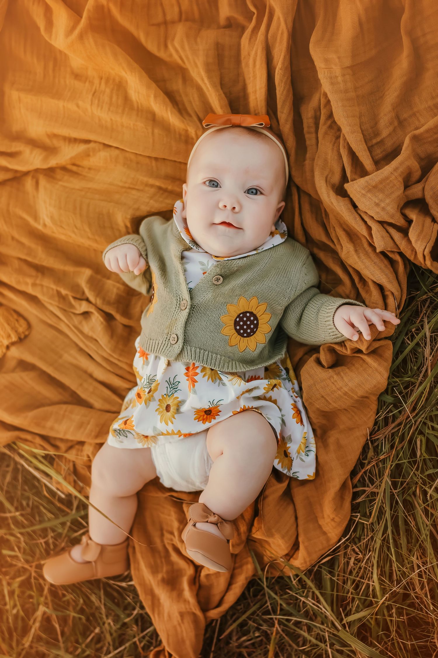 Baby wearing floral dress and cardigan lying on orange blanket outdoors.