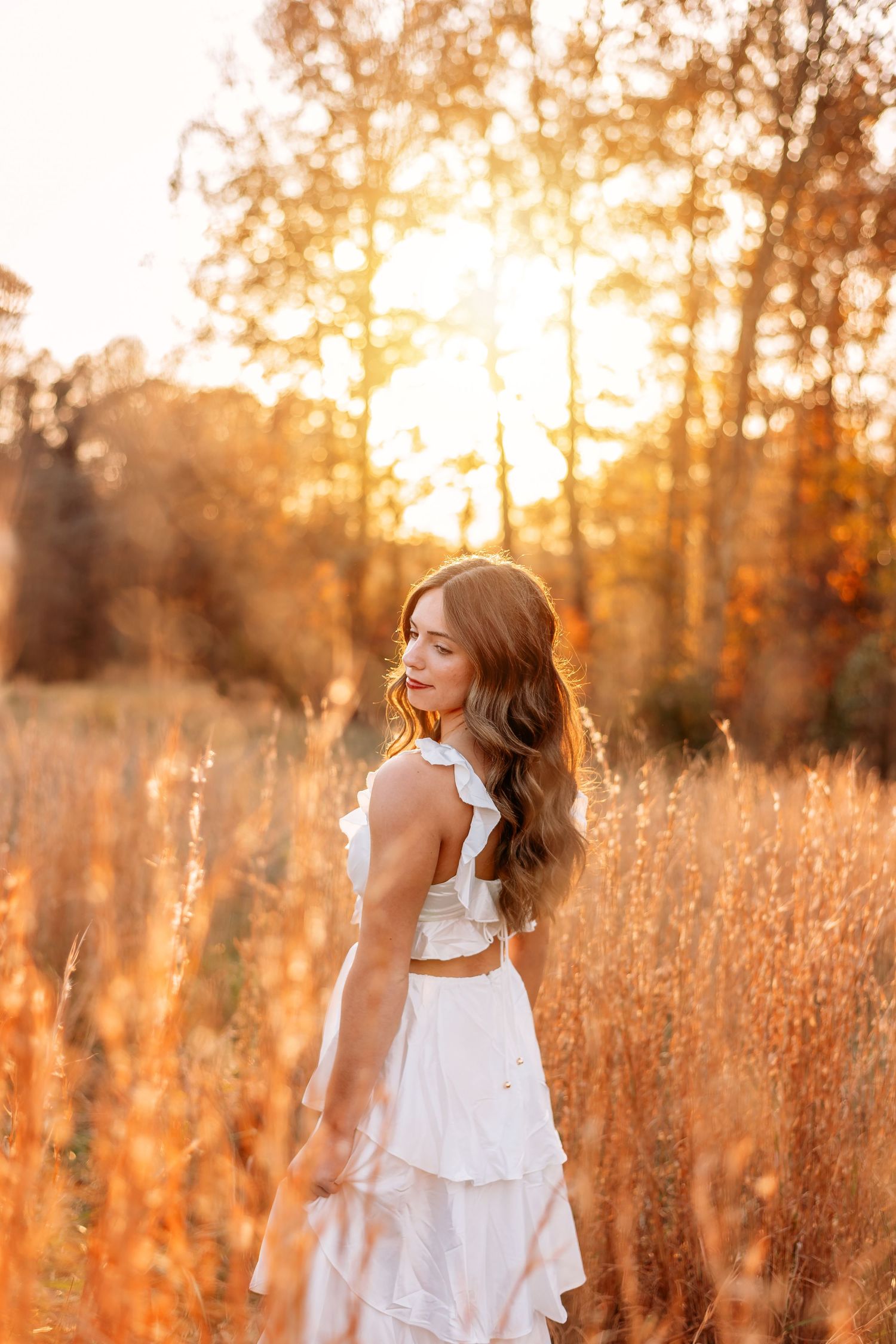 A woman in a white dress stands in a golden field at sunset with sunlit autumn trees in the background.