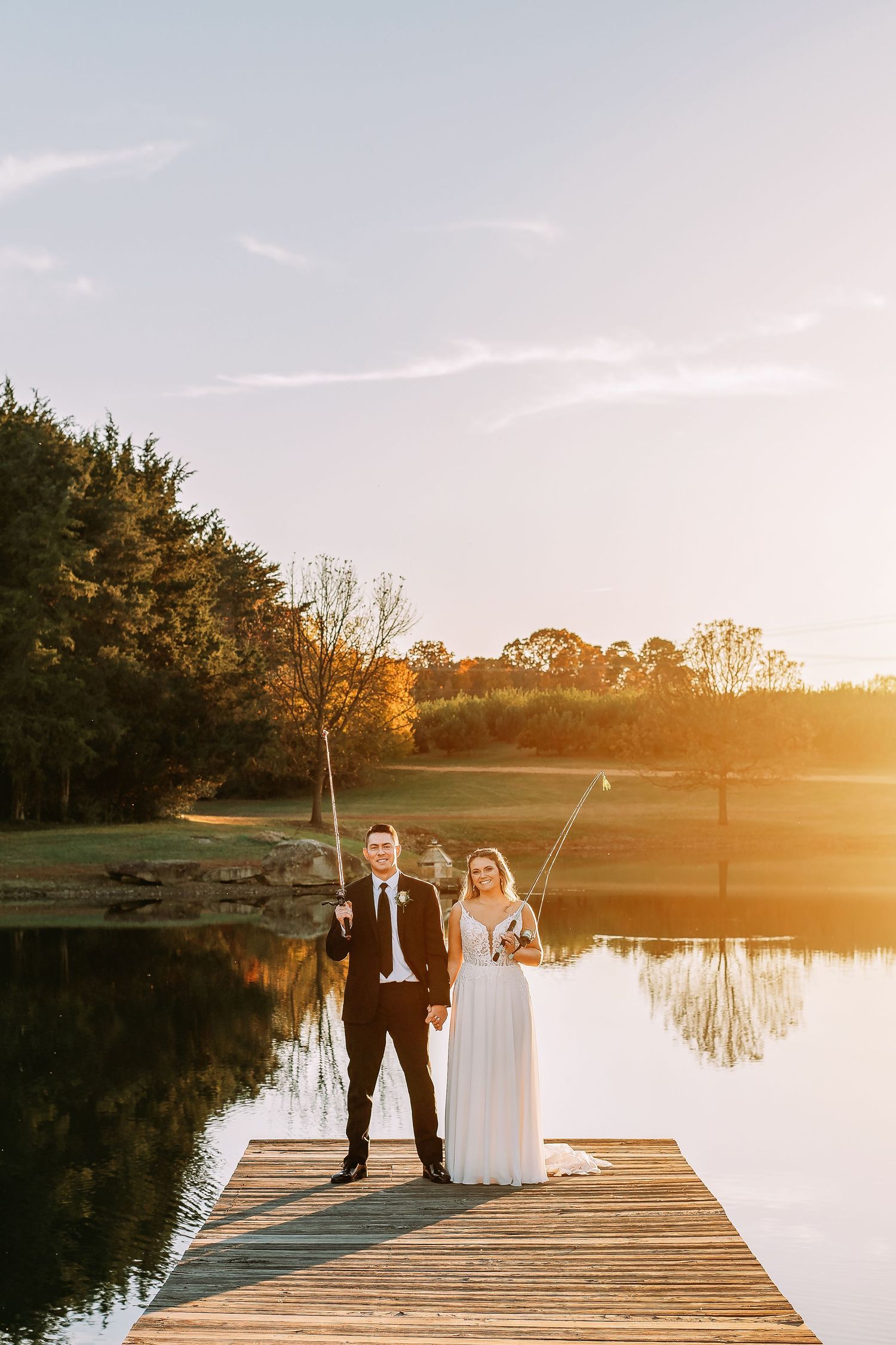 Bride and groom holding fishing rods on a wooden dock at golden sunset with calm lake and trees reflecting in water.