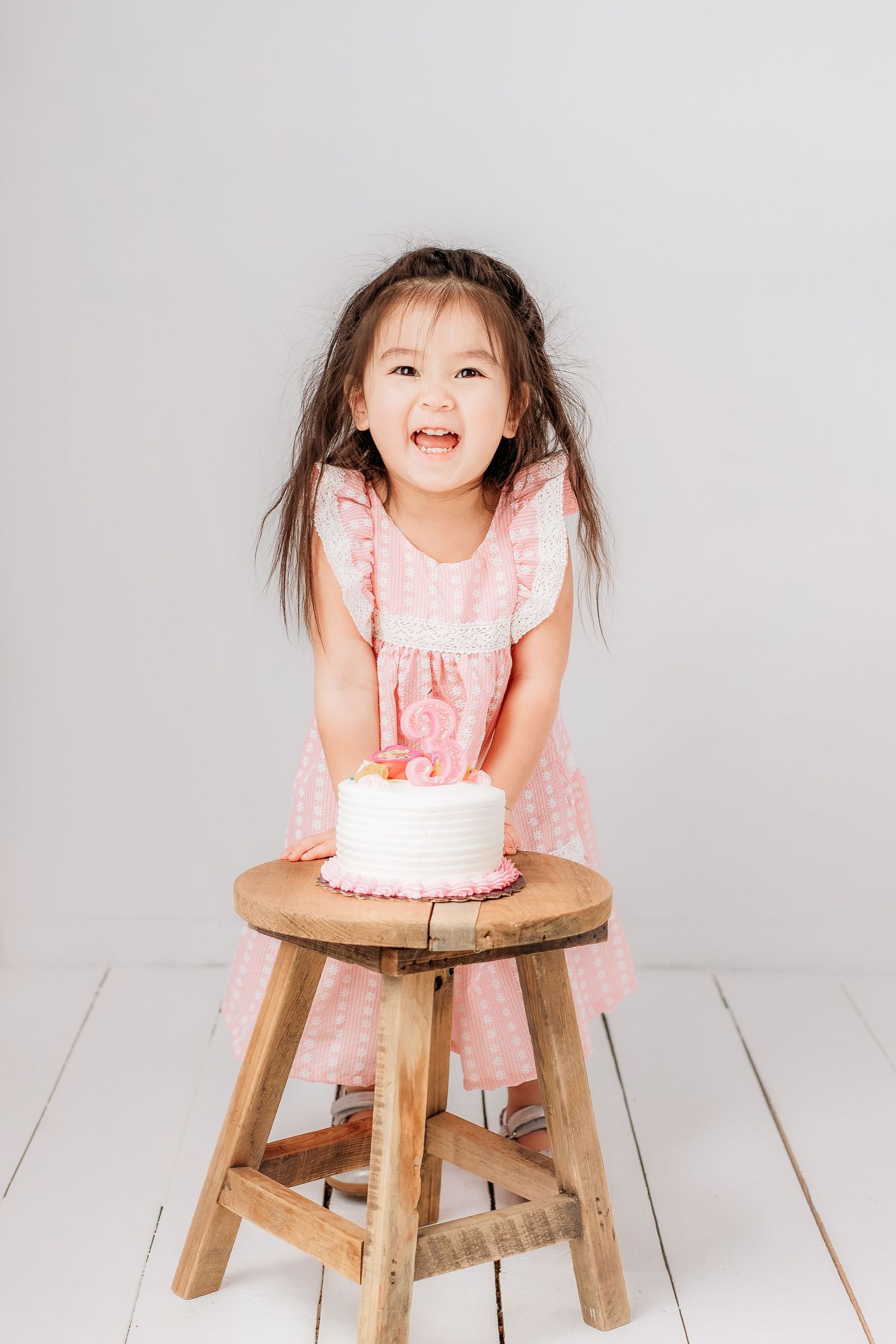 A joyful child in a pink ruffled dress stands at a wooden stool with a white birthday cake in a bright studio setting.