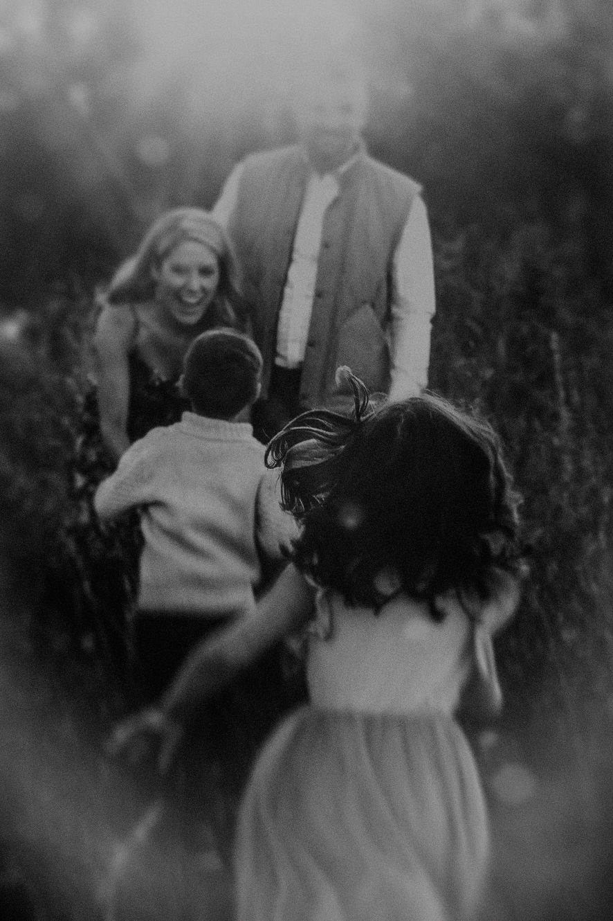 Black and white photo of two children running toward their laughing parents in a sunlit outdoor field.