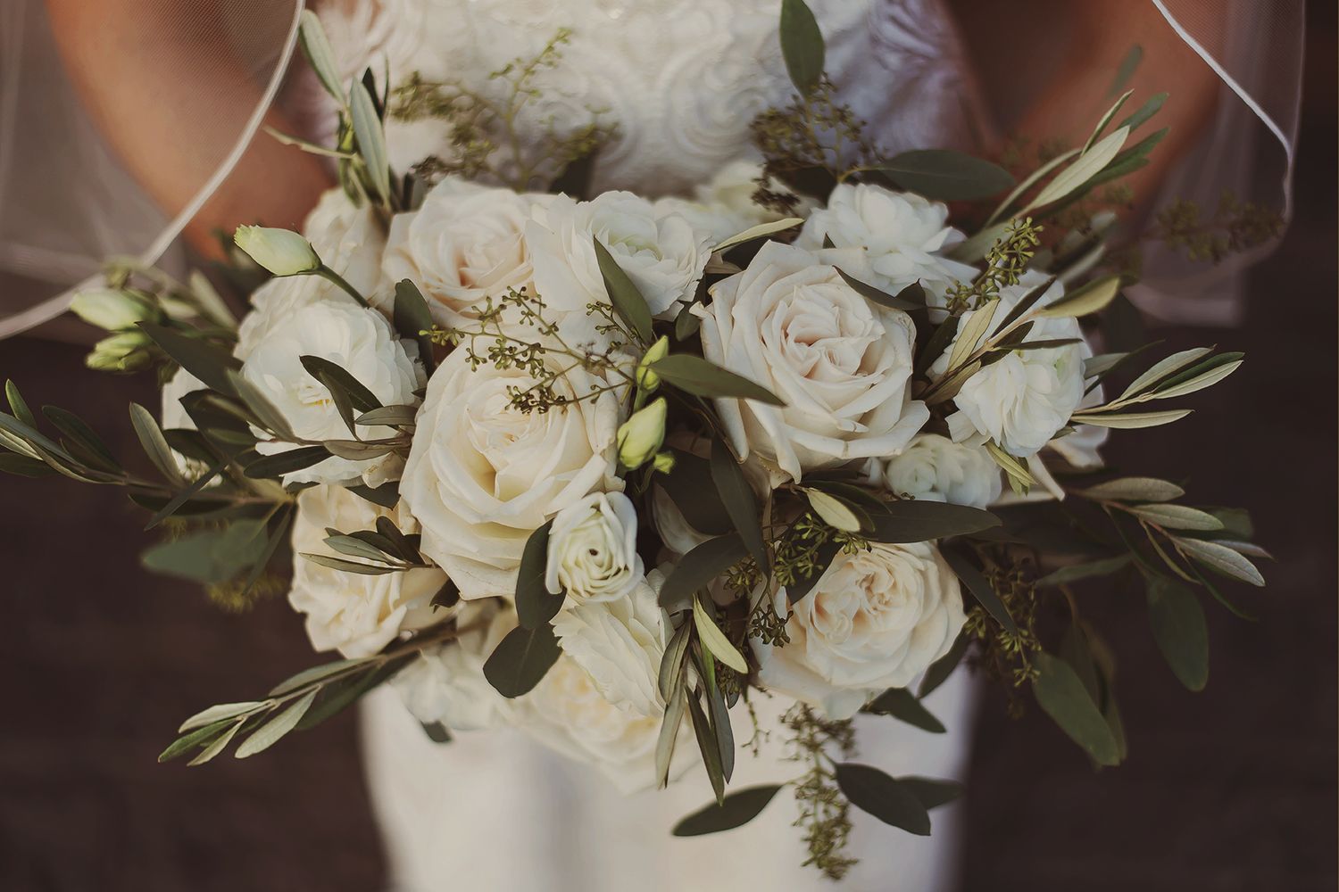 Bride holding a lush white rose and greenery wedding bouquet with olive branches and delicate blooms.