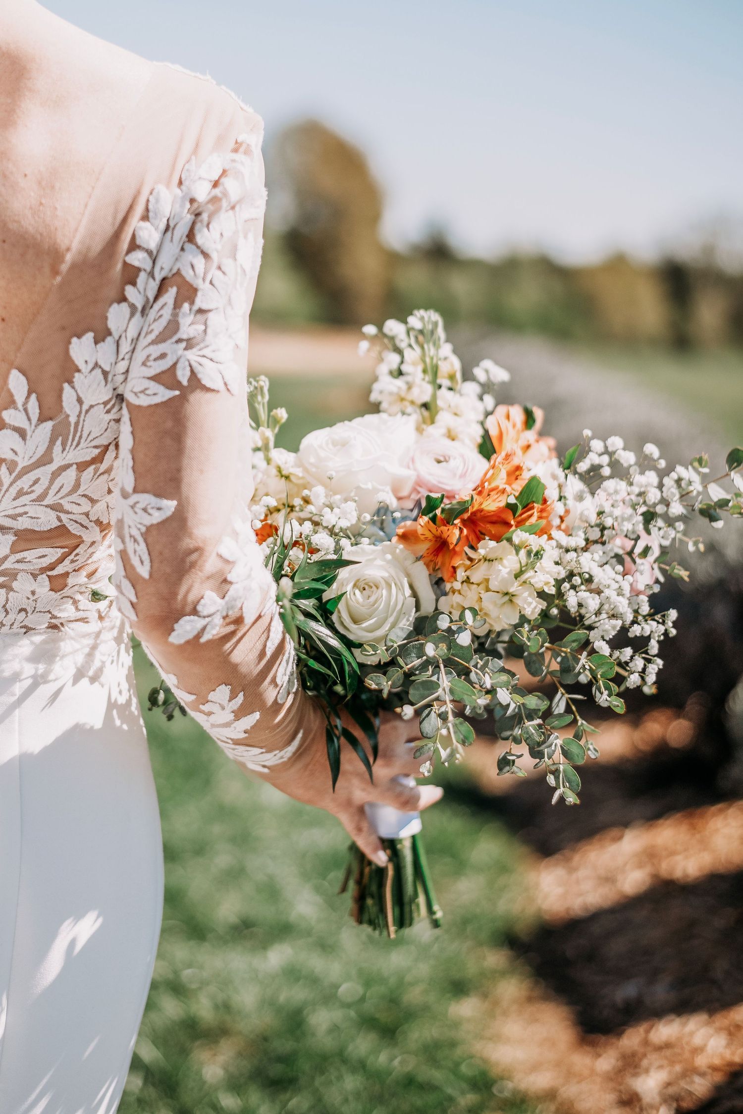 Bride in lace wedding gown holding a bouquet of white roses, orange alstroemeria, and greenery outdoors.