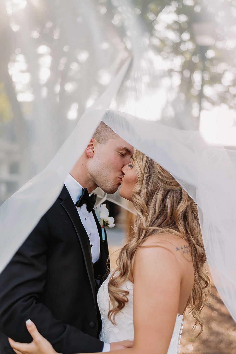 Bride and groom sharing a romantic kiss under a flowing white veil outdoors, groom in black tuxedo, bride in white gown.