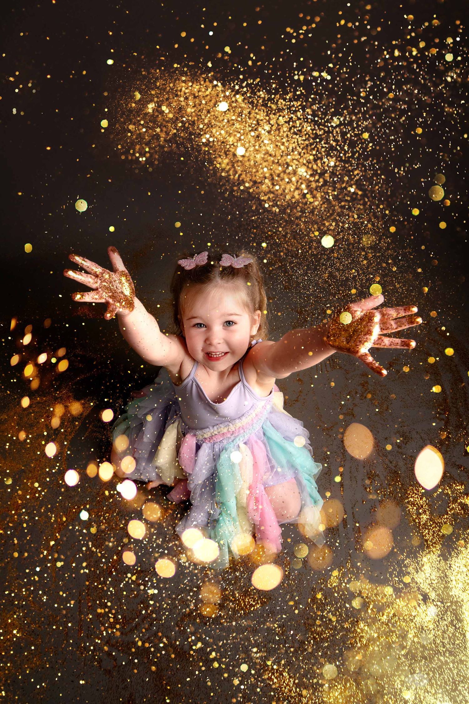 Magical portrait with glittering gold sparkles and bokeh effects surrounding a joyful child in a pastel tulle dress.