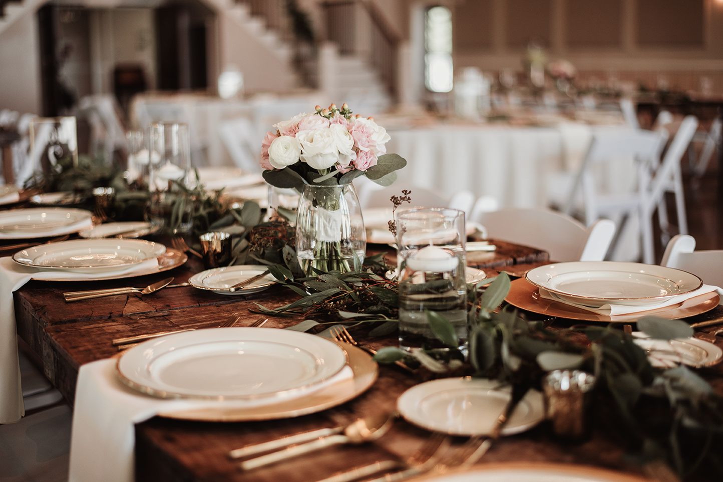 Elegant rustic wedding reception table with greenery runner, white roses, gold chargers, and candles on a wooden farm table.