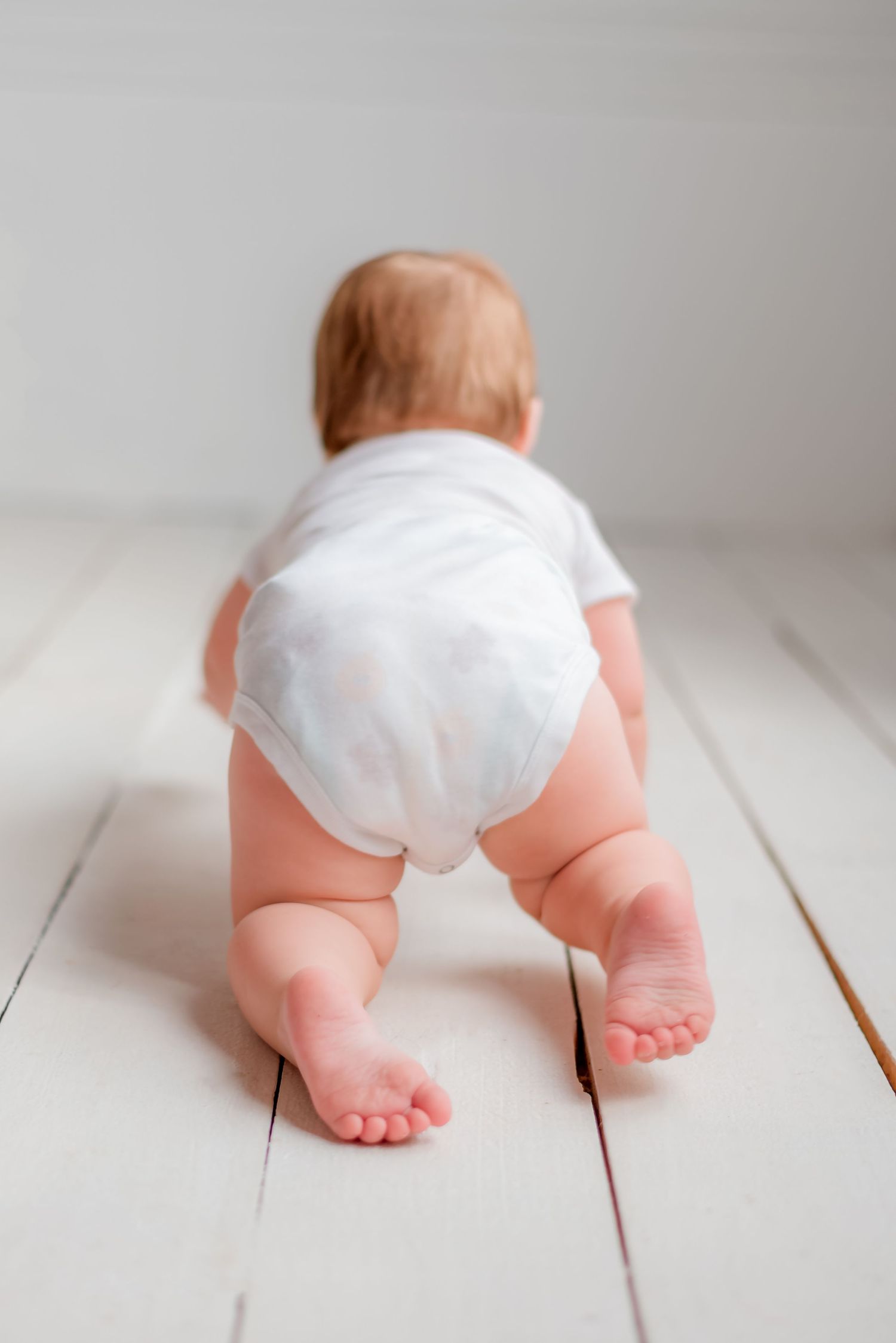 Baby wearing white diaper crawls on wooden floor showing tiny feet and chubby legs.