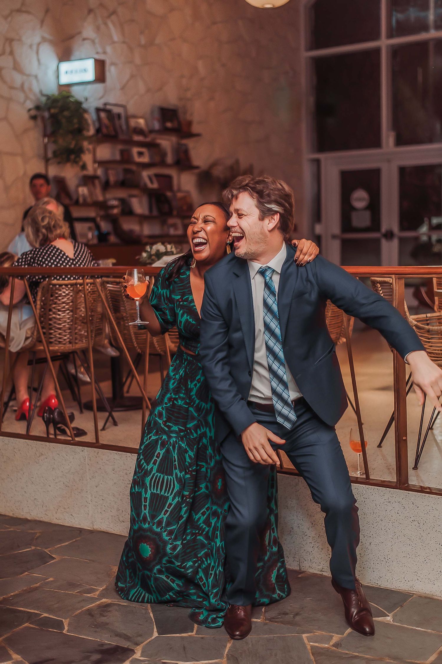 A laughing couple in elegant attire enjoy a lively moment at a restaurant, woman in green dress holding a cocktail.