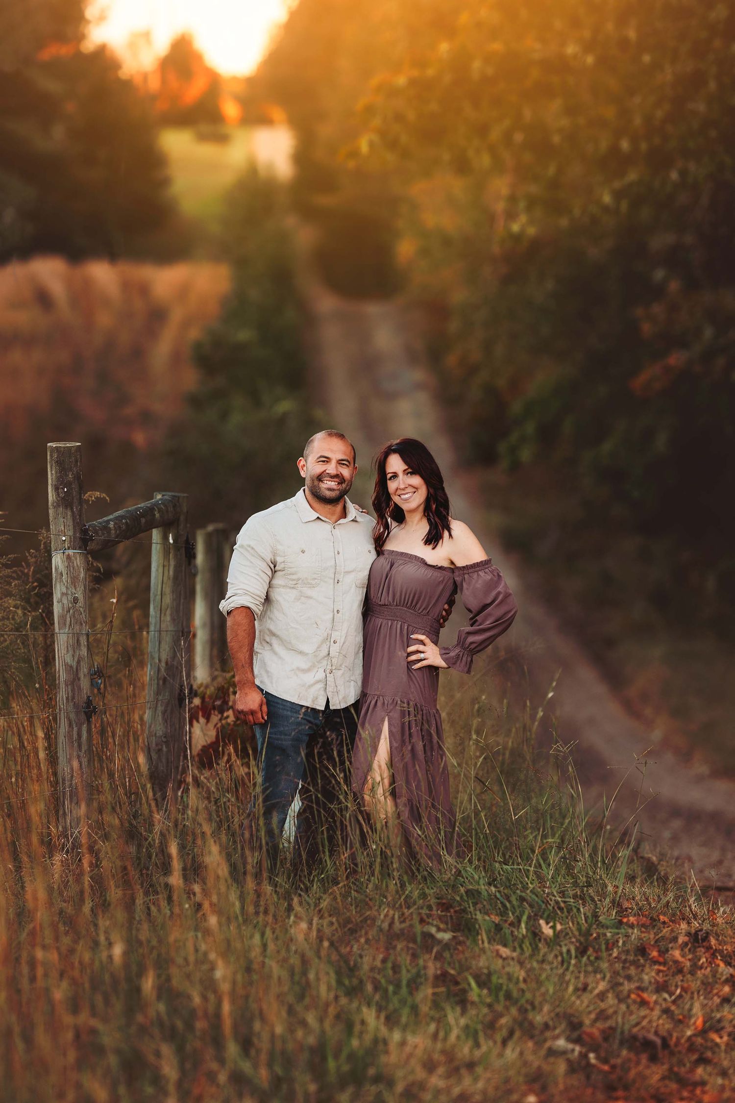 Couple posing by rustic fence on dirt road at golden sunset, woman in purple off-shoulder dress, man in white shirt.