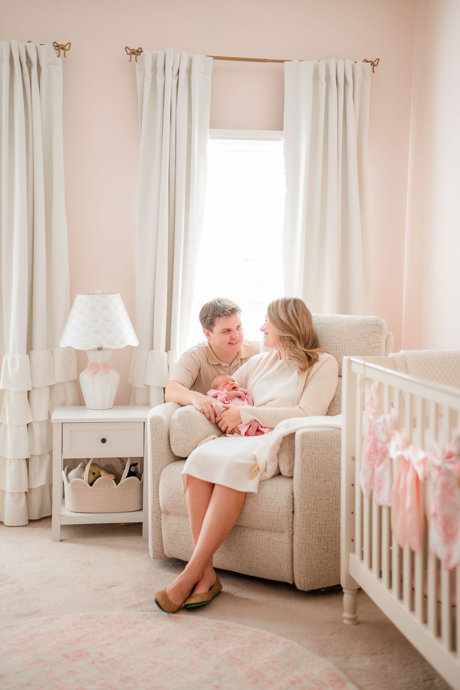 A cozy nursery scene with white curtains and a comfortable armchair next to a white crib.