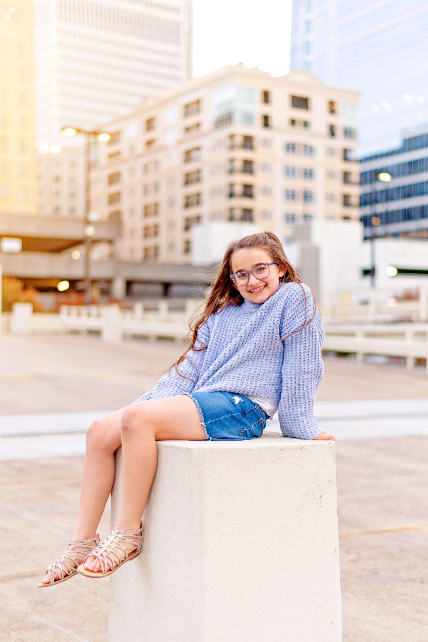 Person in blue sweater sitting on concrete block with city buildings in background.