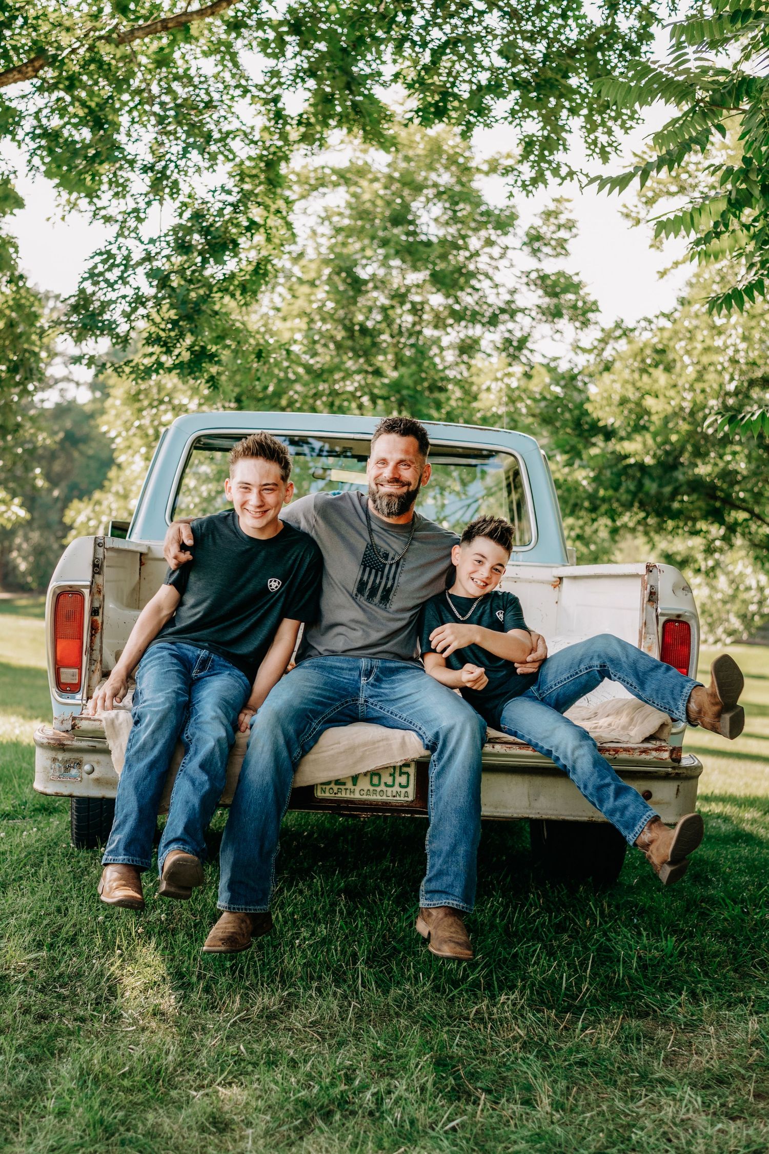 A man and two boys sit smiling on the tailgate of a vintage pickup truck outdoors surrounded by green trees.