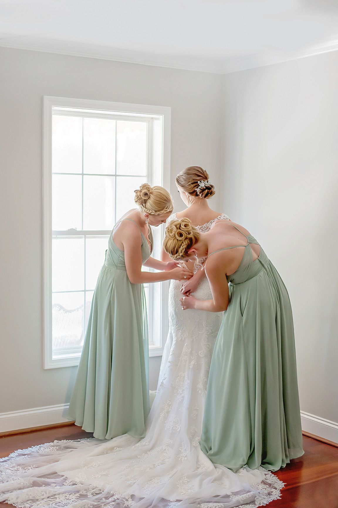 Two bridesmaids in sage green gowns help a bride in white lace dress near a bright window before her wedding ceremony.