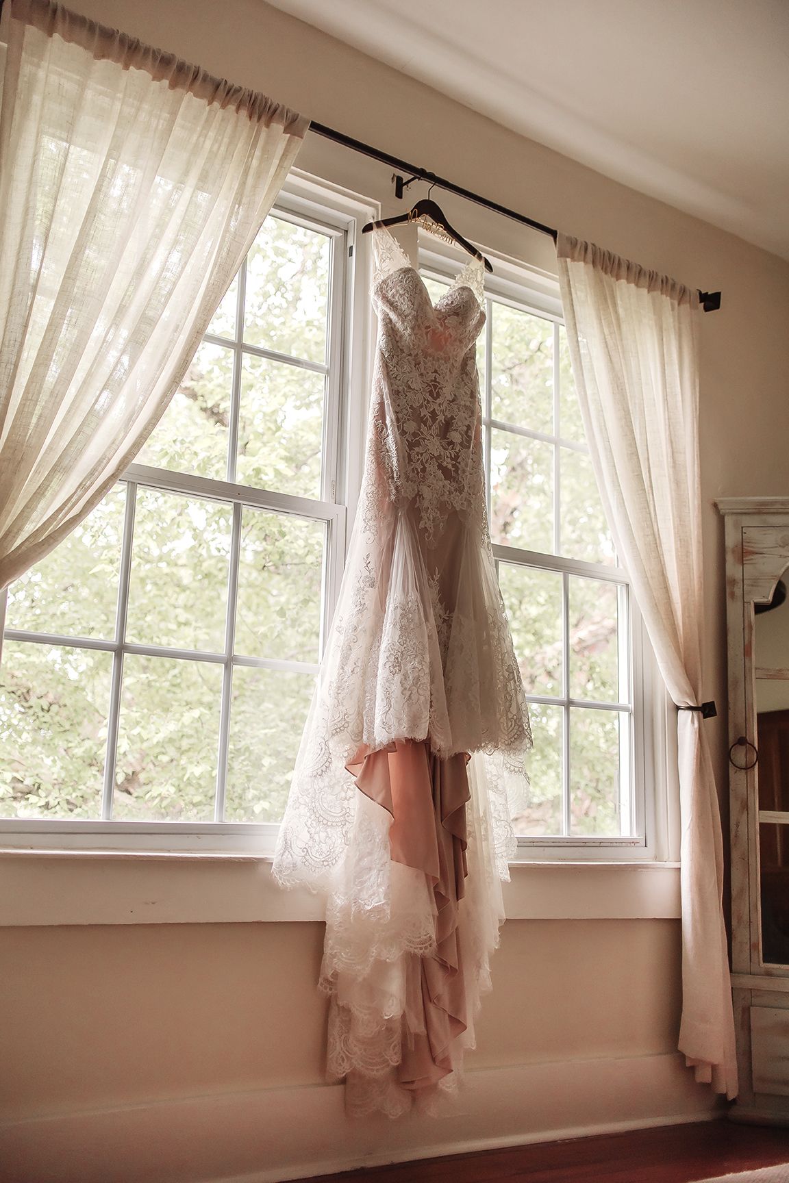 Elegant lace mermaid wedding dress hanging on a curtain rod in front of a sunlit window with sheer drapes.