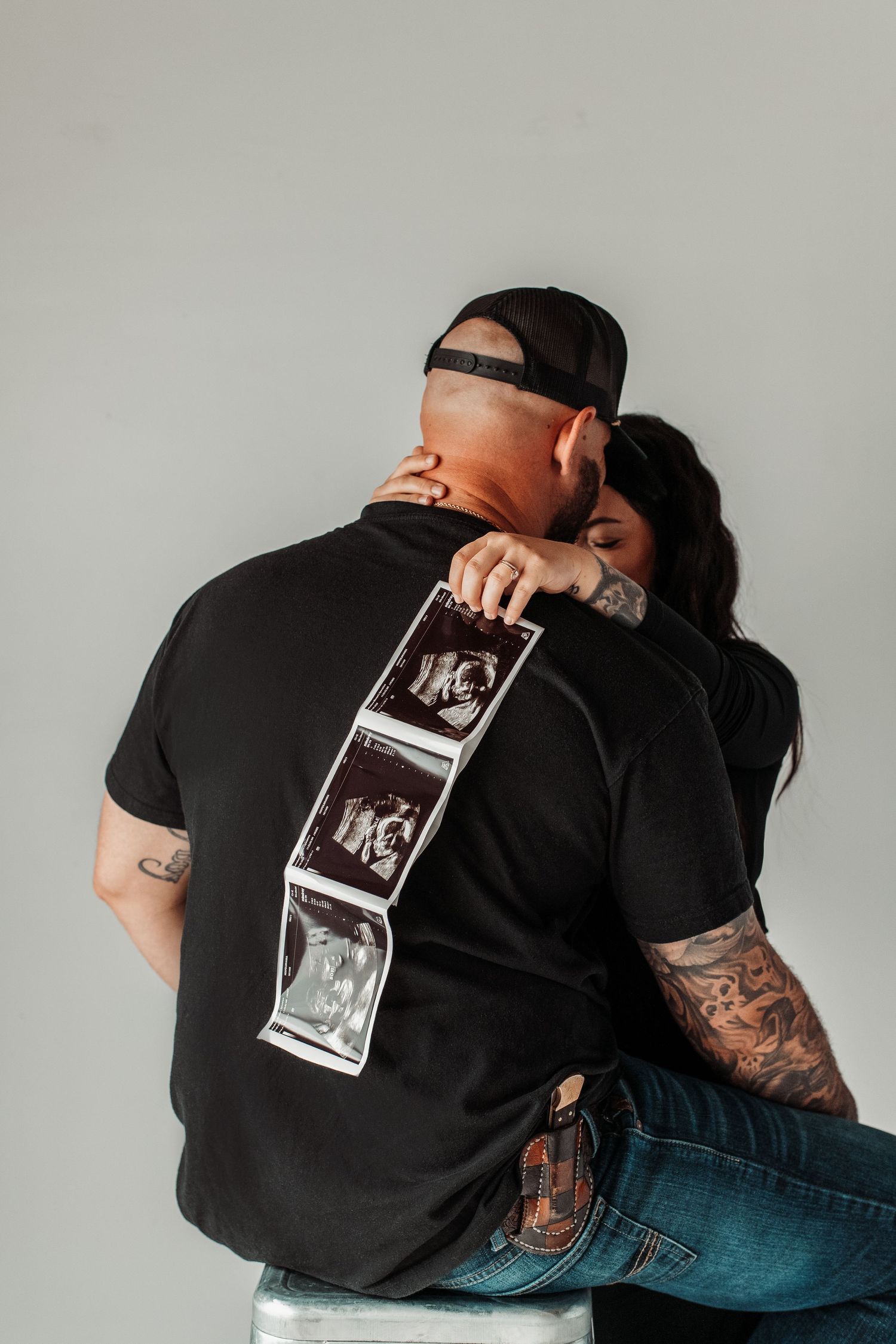 Intimate couple pose with ultrasound photos displayed on person's back wearing black shirt.