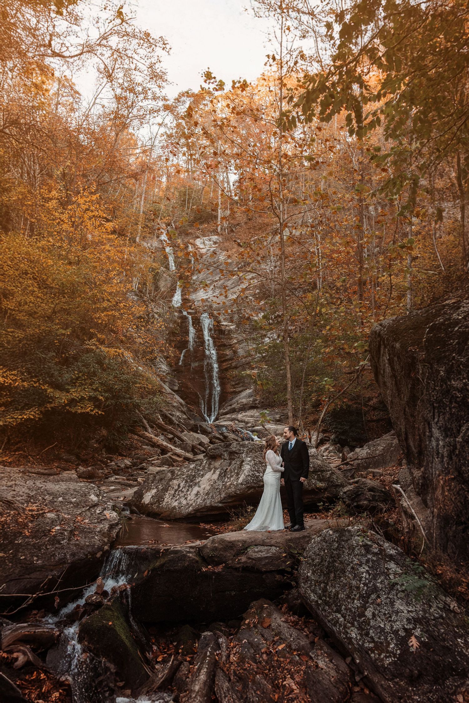 Bride and groom embrace on rocky terrain before a waterfall surrounded by autumn foliage in a forest wedding portrait.