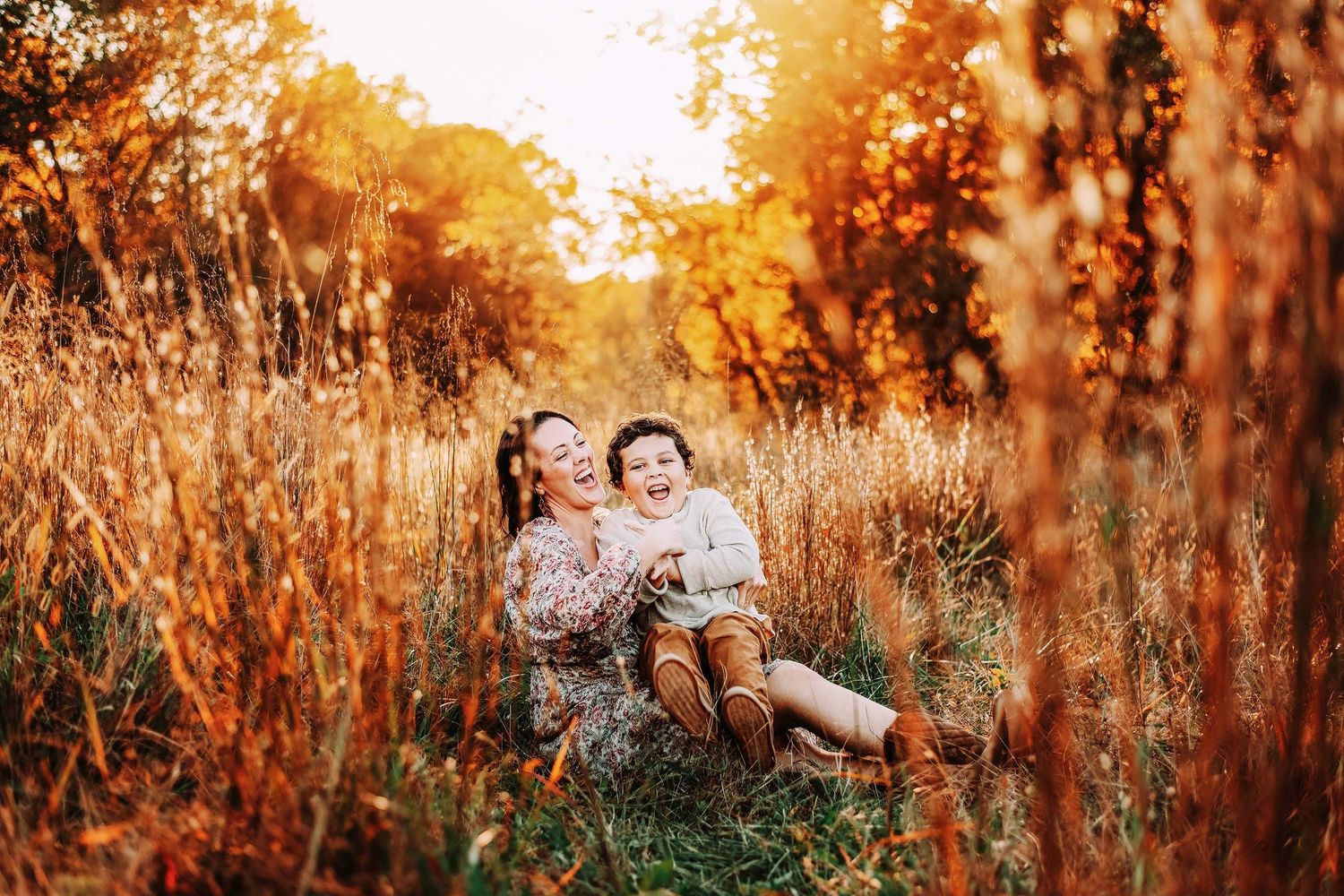 A mother and child laugh joyfully together in a golden autumn field at sunset.