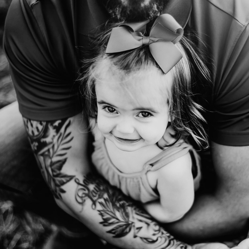 Black and white portrait of a small child wearing a bow, smiling while being held near tattooed arms.