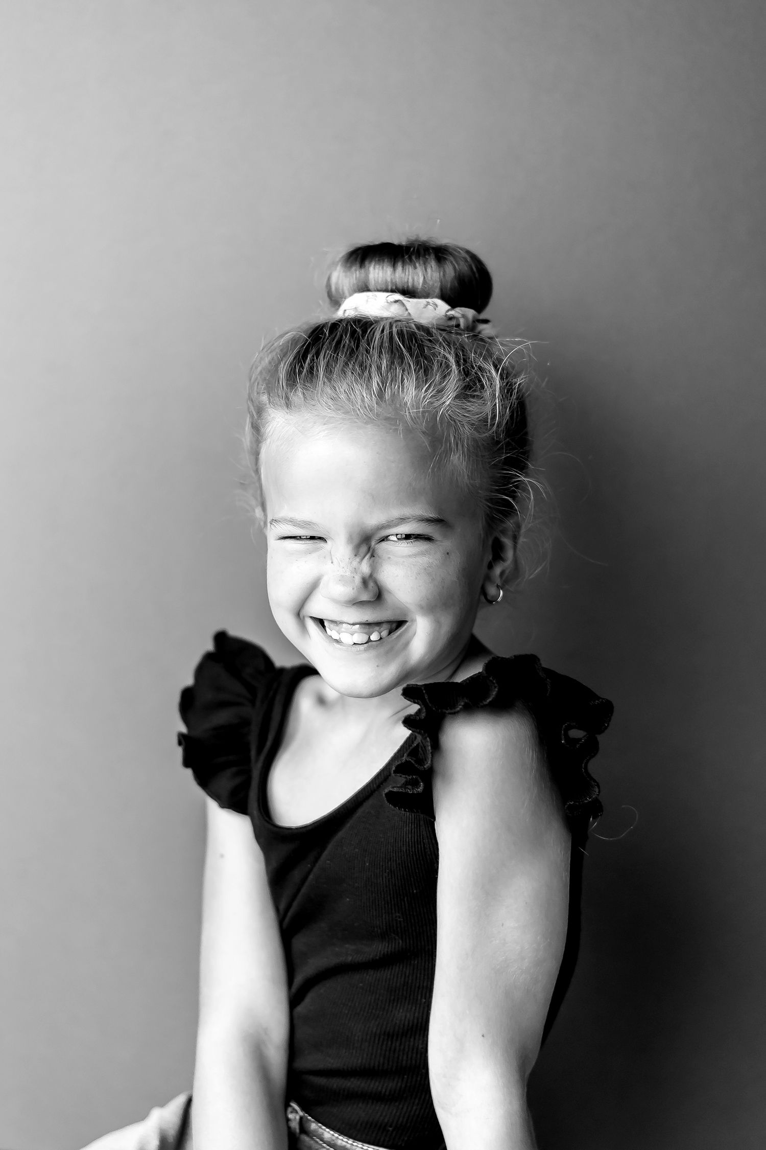 Black and white portrait of a child in a sleeveless ruffled top with hair in a bun showing a bright, joyful smile.