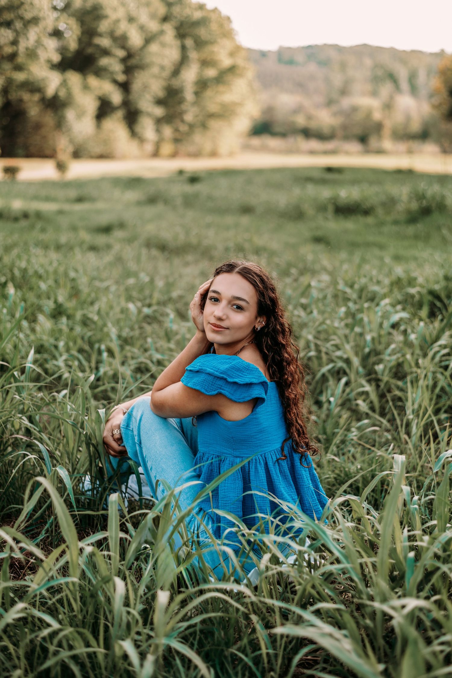 Figure in bright blue dress sitting among tall grass in a sunlit meadow.