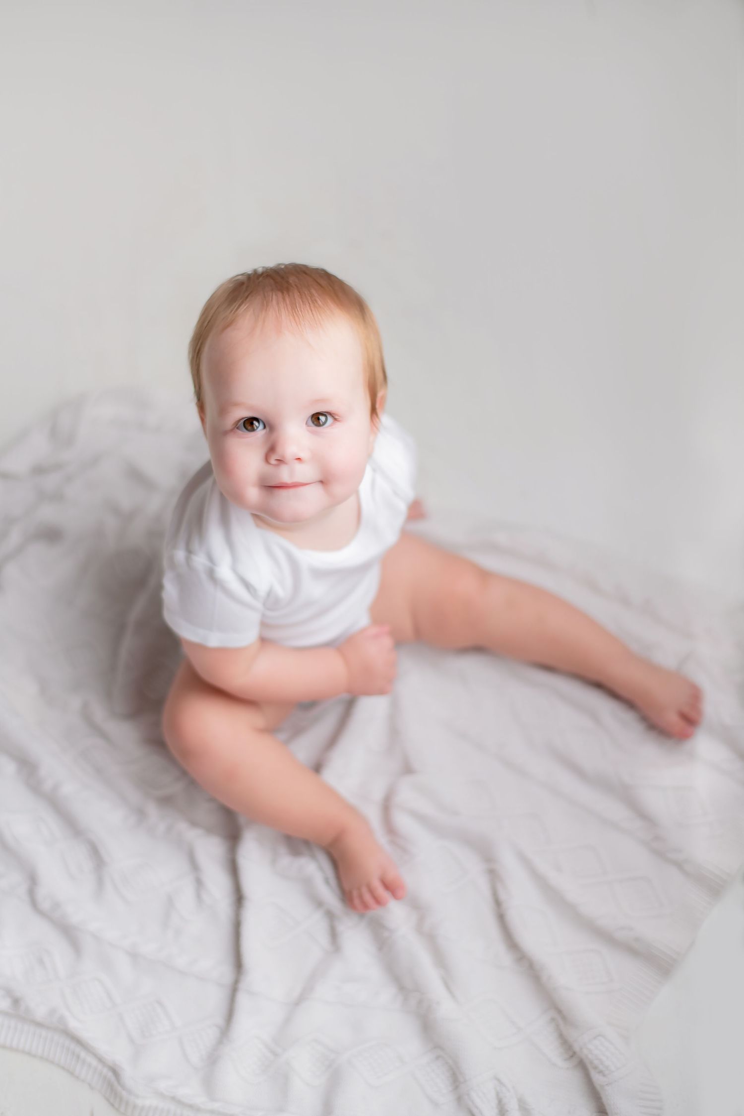 Adorable baby wearing a white onesie sits on soft gray blankets in a minimalist portrait setting.