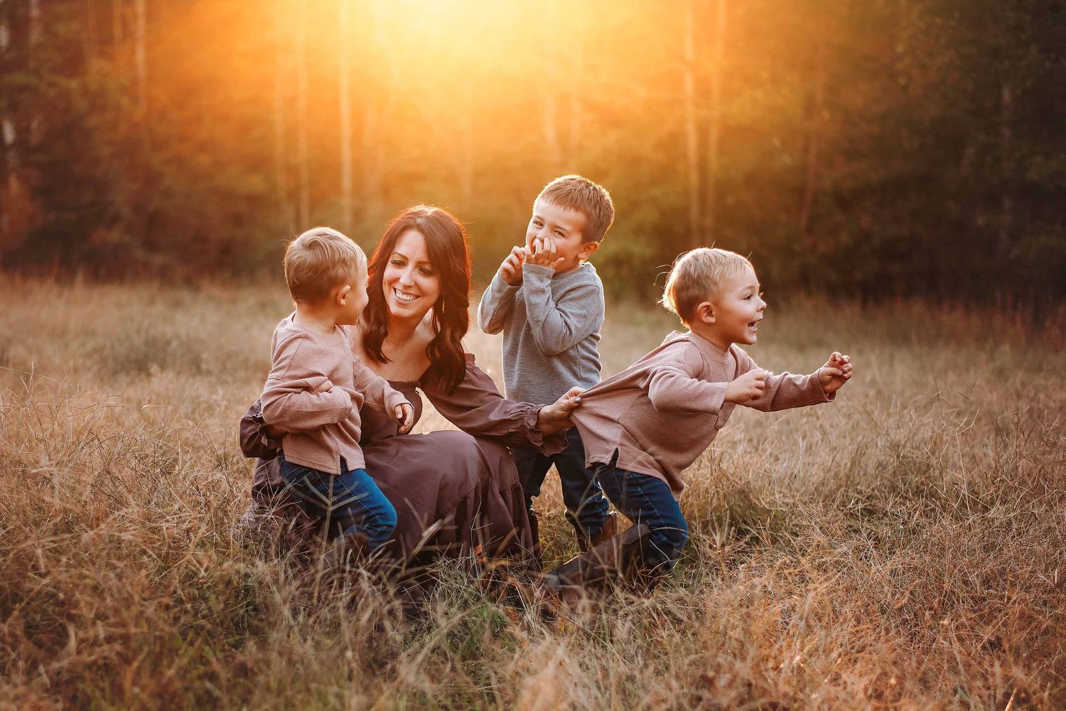 A mother sits in a golden sunlit field with three young boys laughing and playing around her during sunset.