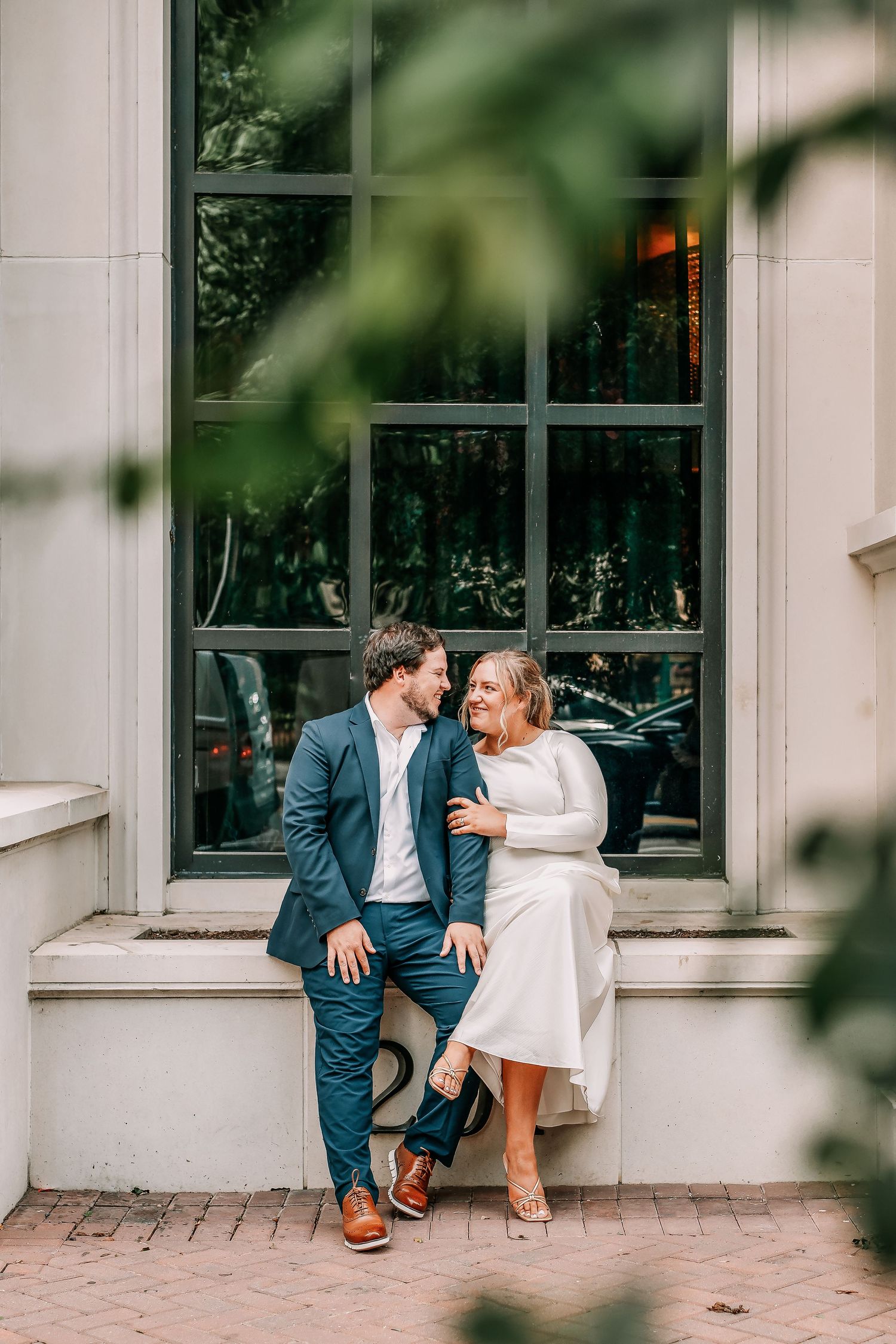 Couple in wedding attire sitting together on a window ledge, sharing a romantic moment outdoors.