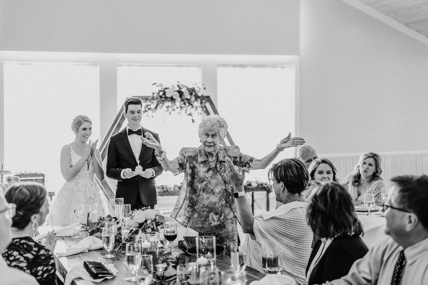 Black and white wedding reception photo of bride and groom with elderly guest dancing joyfully at elegant indoor celebration.