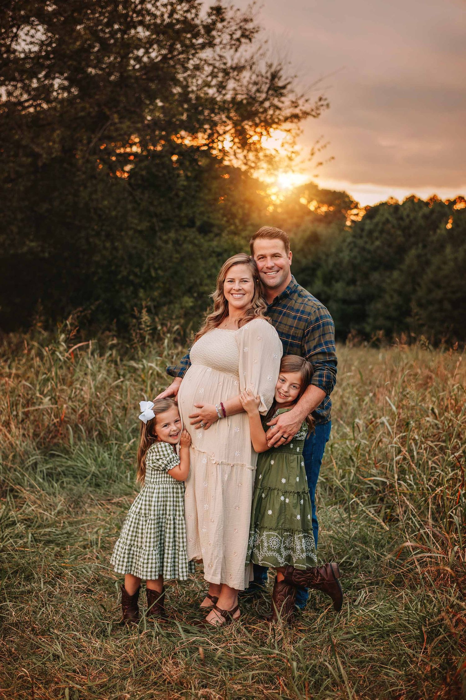 Pregnant woman and man pose outdoors at golden sunset with two young girls in coordinating outfits in a field.