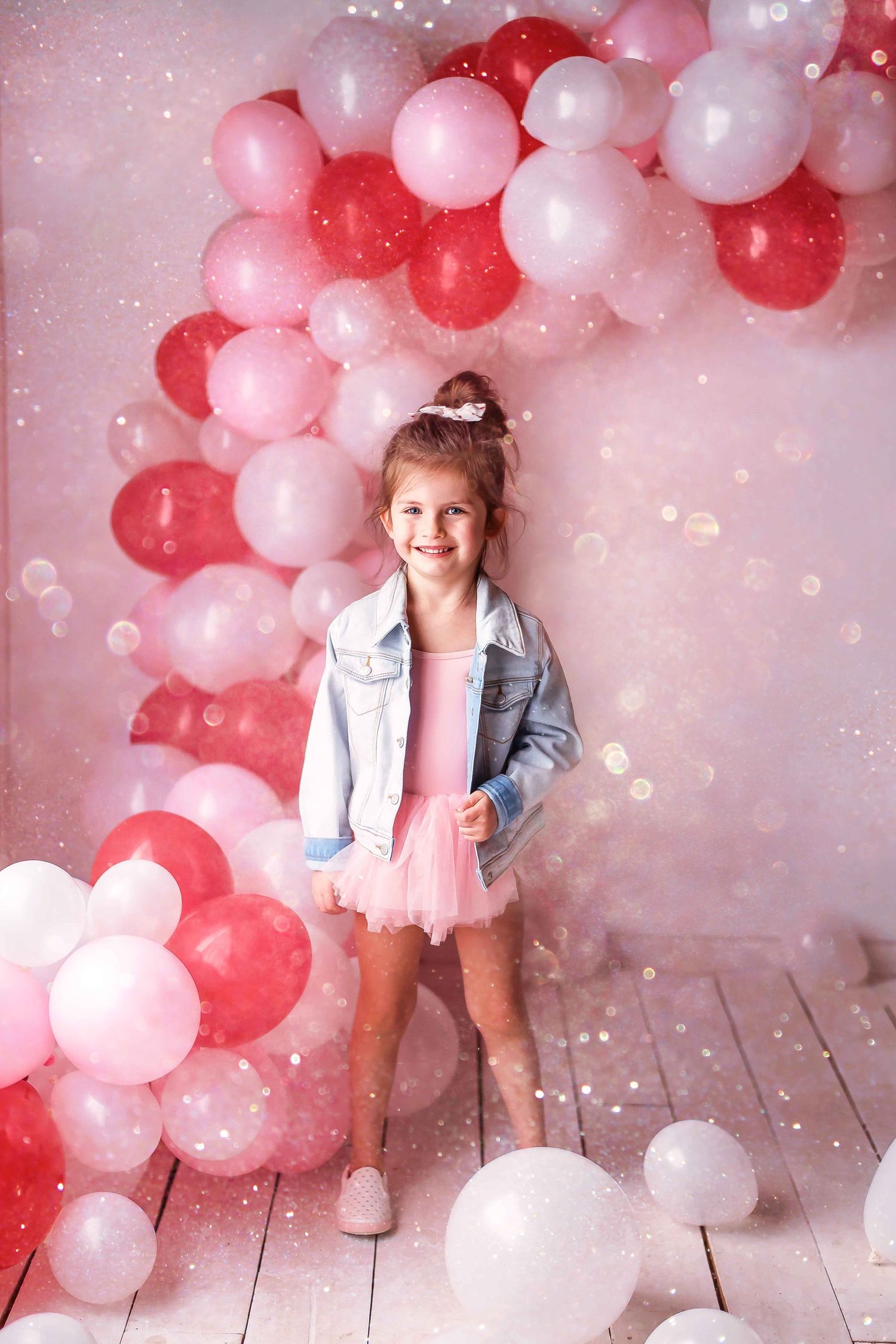 Festive pink and red balloon arch decorates a wooden floor setting with sparkly bokeh effects in the background.