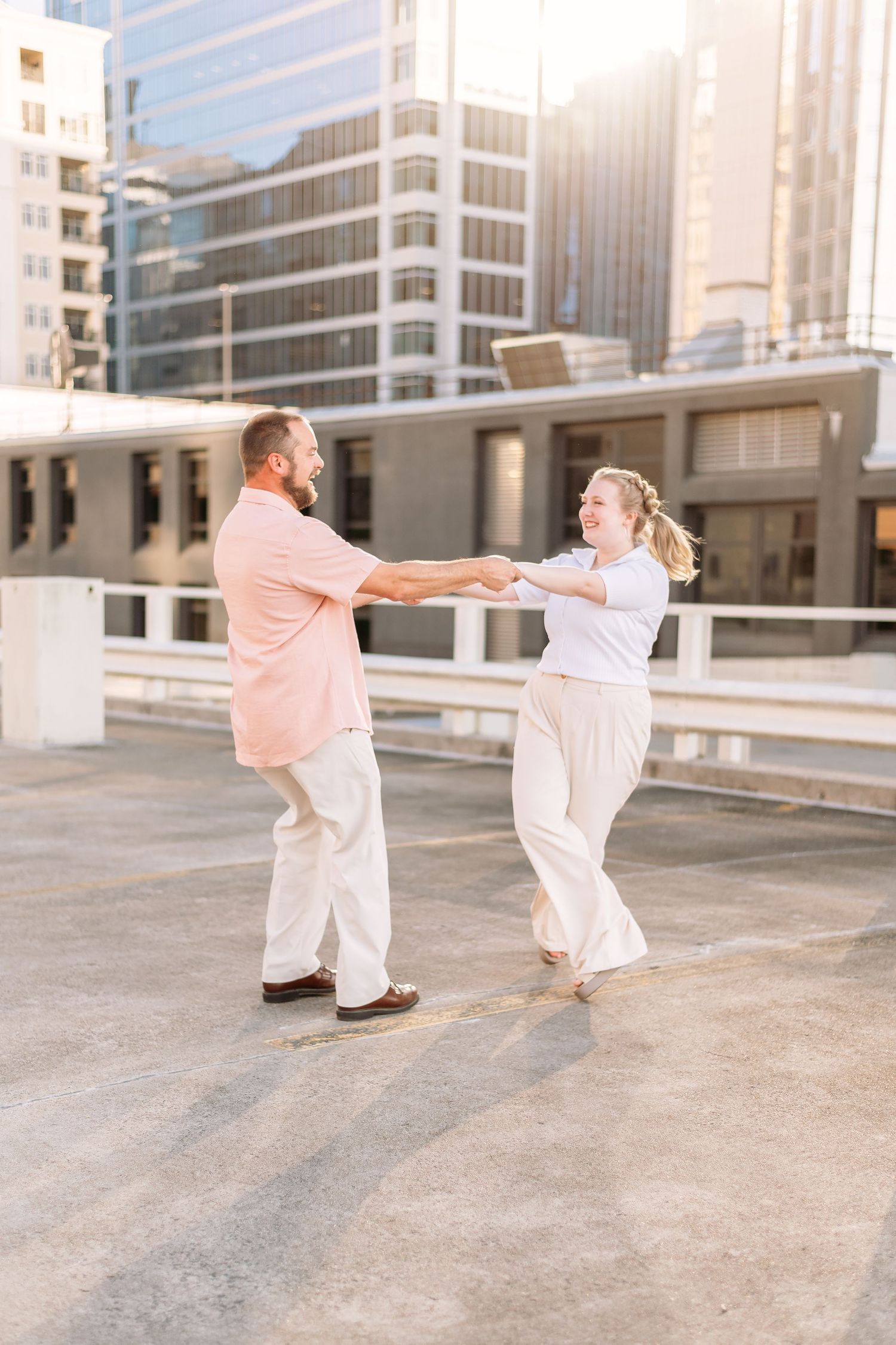 Playful couple in white and pink outfits dancing together on an urban rooftop with city buildings in the background.
