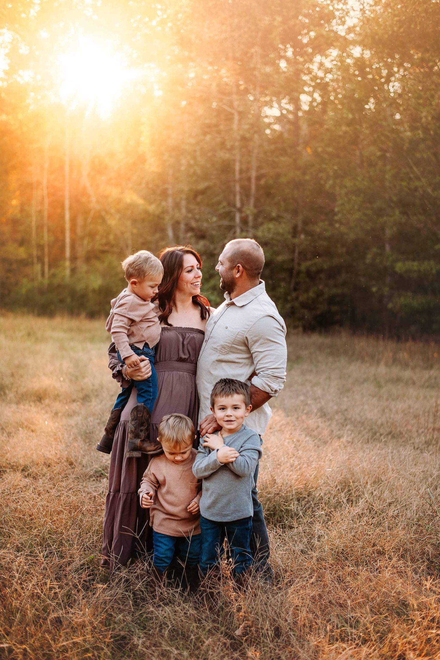 Family of five posing in golden sunlit field at sunset, parents holding children among tall grass with trees in background.