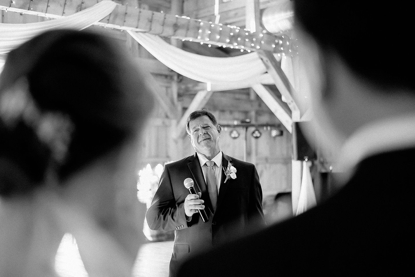 Black and white photo of a man giving a wedding speech at a rustic barn reception venue.