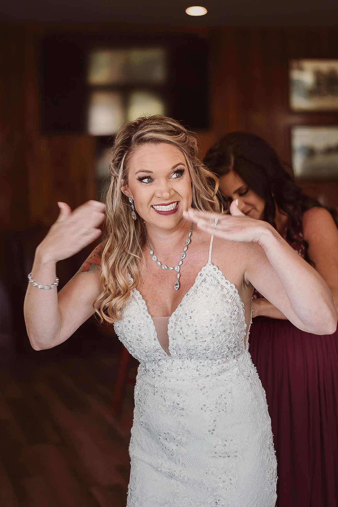 Bride in beaded white gown smiles as bridesmaid in burgundy dress fastens her necklace before wedding ceremony.
