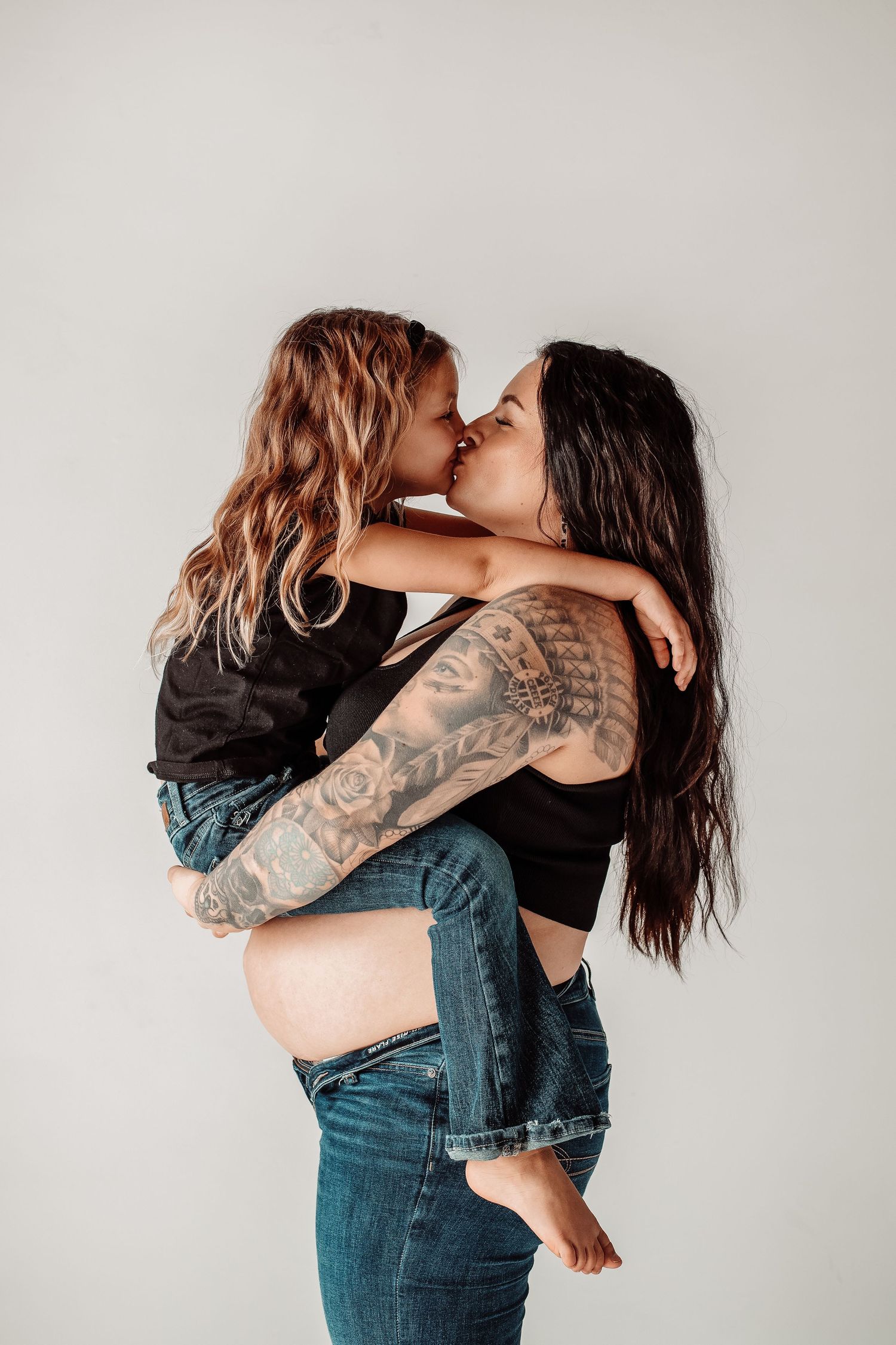 Tattooed pregnant woman holding and kissing a young girl in jeans against a white background.
