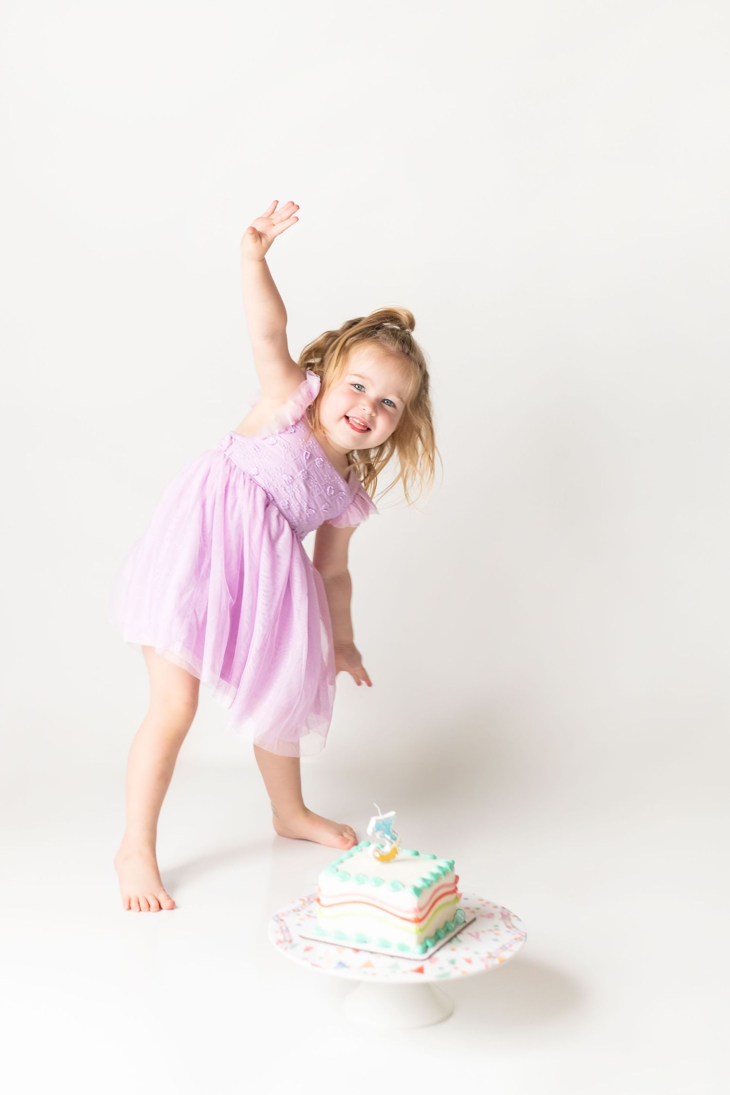 Child in pink dress jumping playfully next to a colorful birthday cake on white cake stand against plain background.