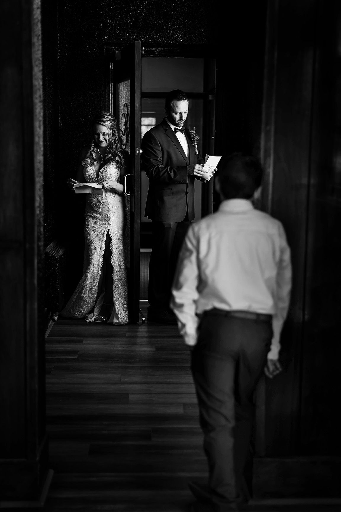 Black and white wedding photo of bride and groom in doorway reading vows while guest stands in foreground.