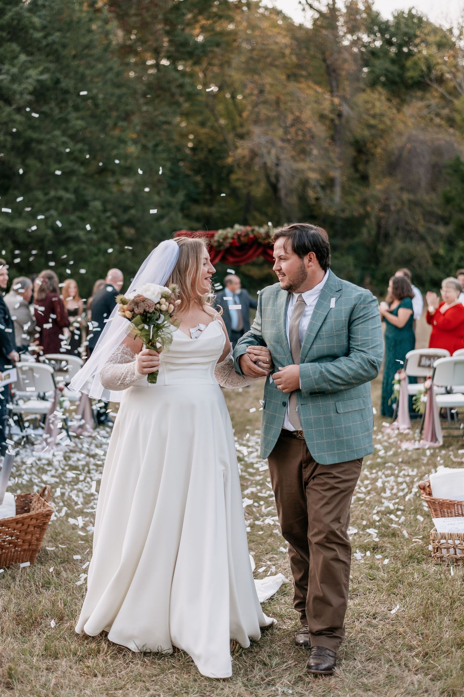 Bride and groom walk down outdoor aisle as guests toss white confetti petals at fall wedding ceremony.