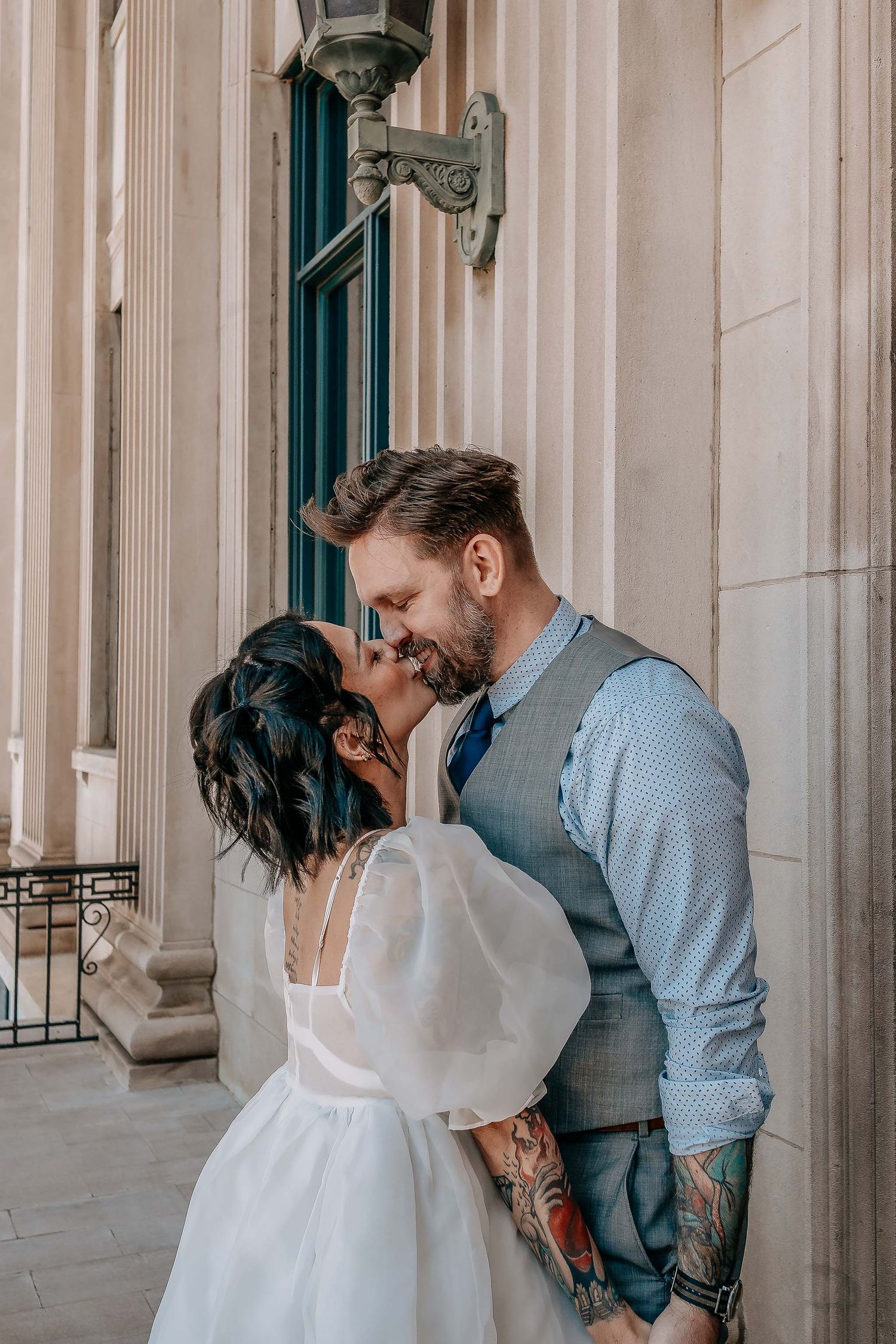 Bride and groom sharing a romantic kiss outside a grand building with stone columns, groom wearing a gray vest with tattoos.