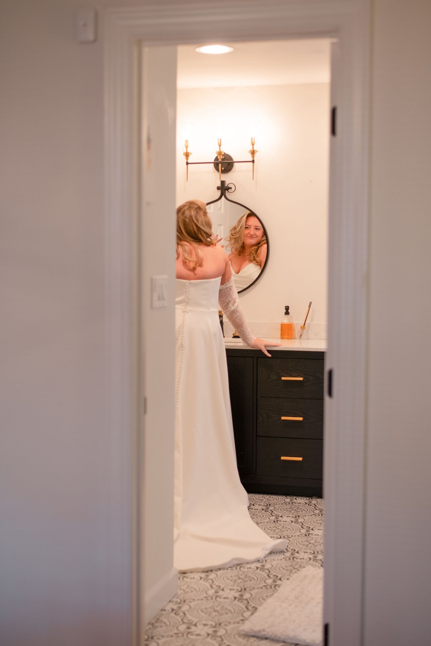 Bride in white wedding gown preparing in elegant bathroom with dark vanity, round mirror, and warm wall sconces.