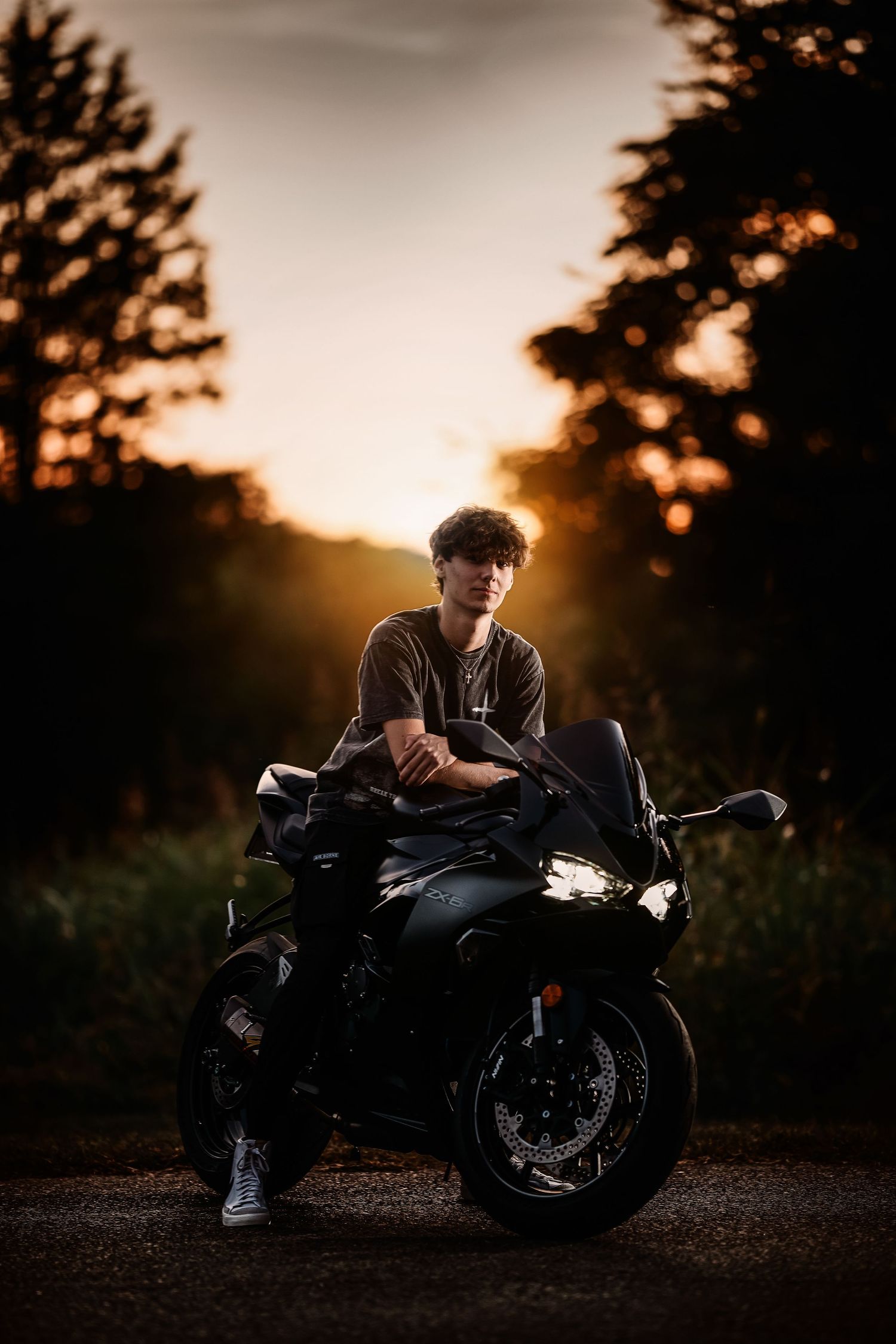 Silhouette of a person sitting on a black motorcycle against a golden sunset with trees in the background.