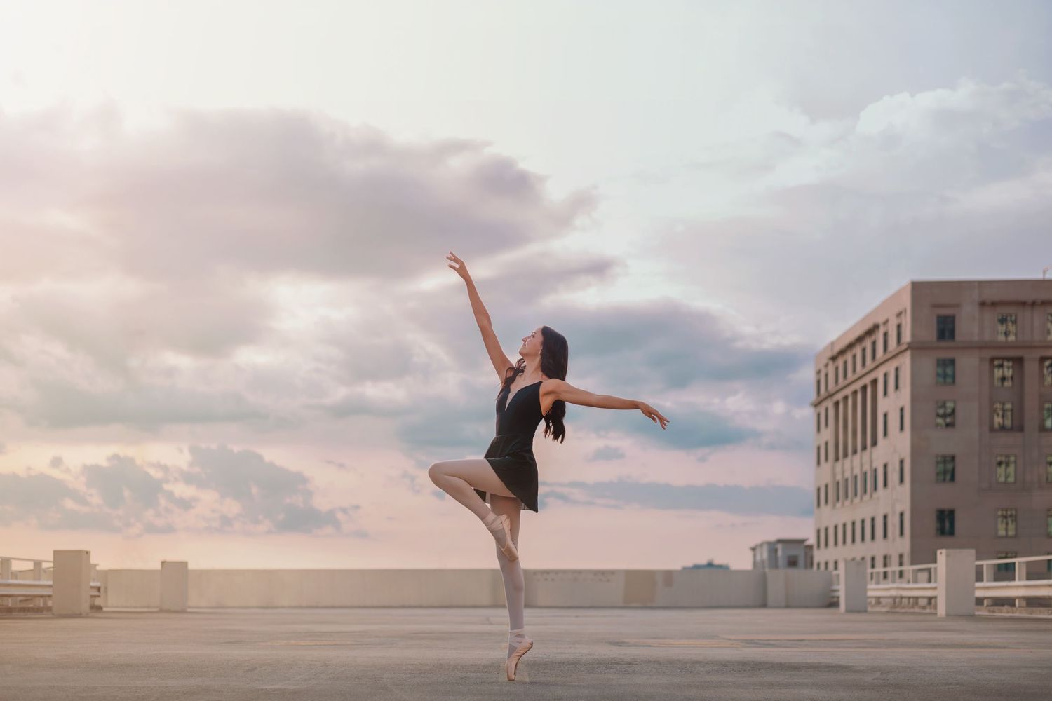 Ballet dancer in a black dress performing an elegant pose on a rooftop at sunset.