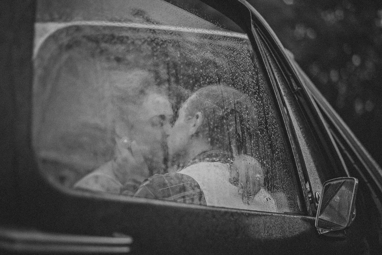 Black and white photo of a couple kissing inside a car with rain droplets on the window.